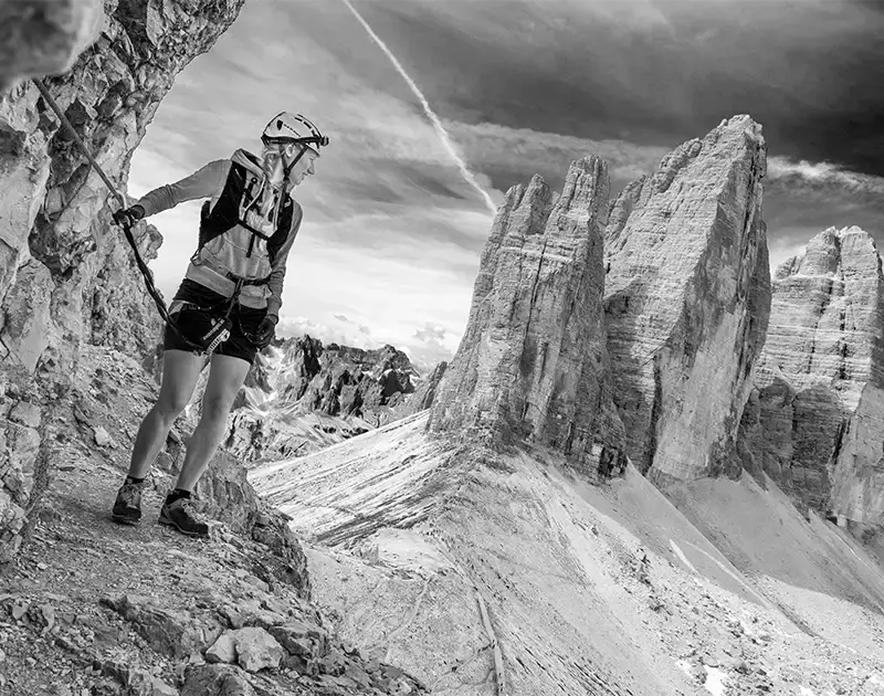 Frau mit Helm und Seilsicherung steht in einem Klettersteig Blick auf die Berge der 3 Zinnen