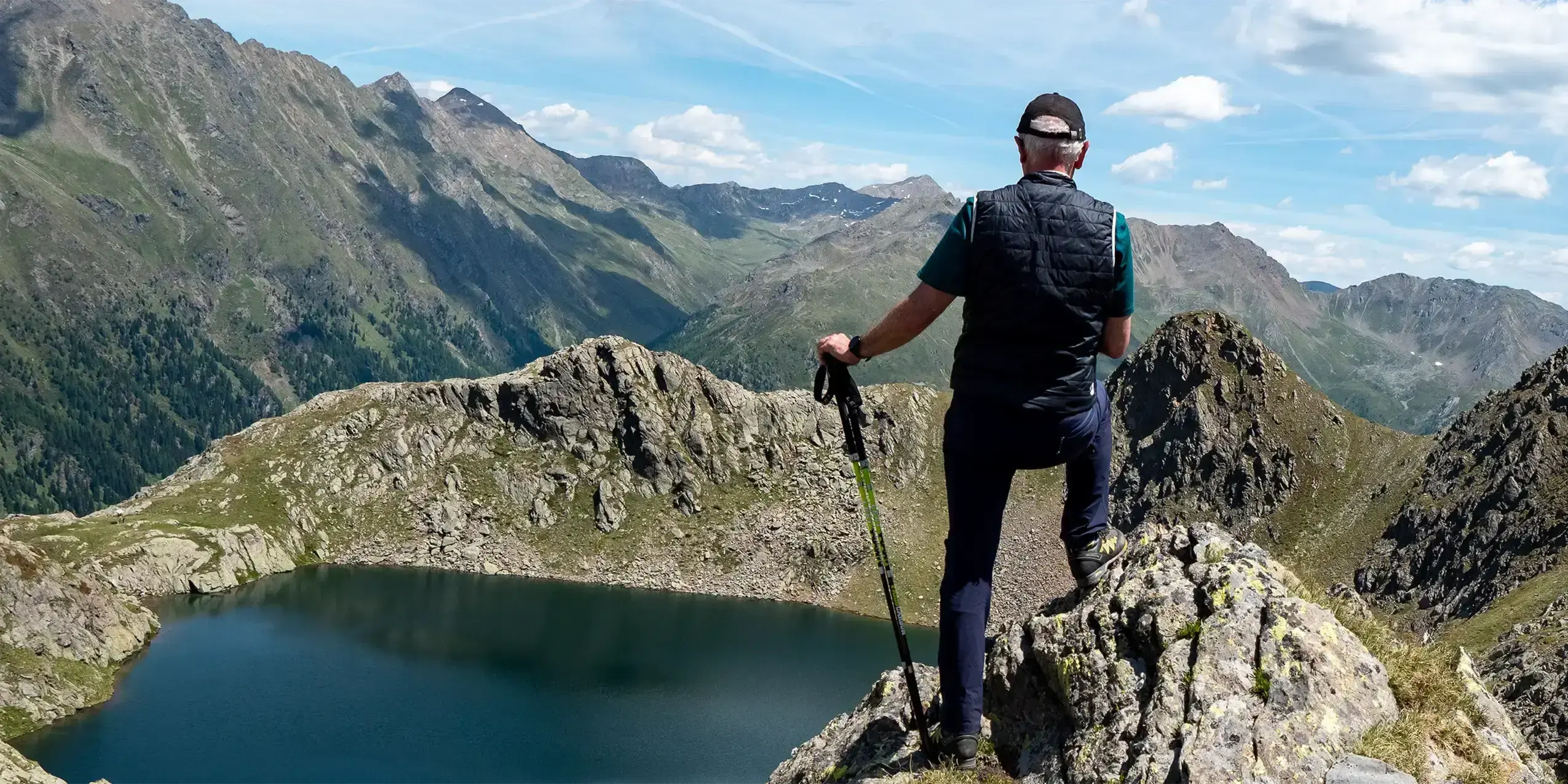 Mann steht im Gebirge oberhalb eines Bergsees und schaut auf die Berge
