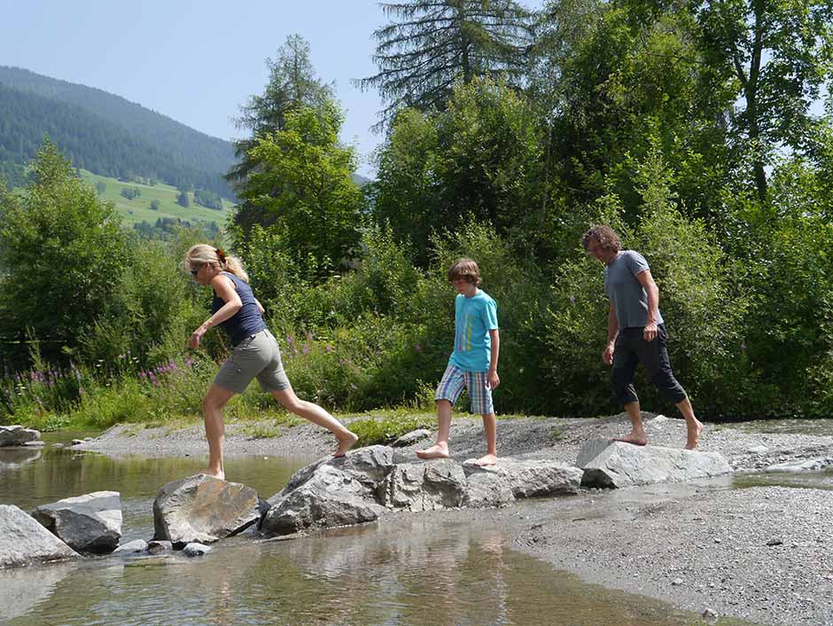 eine Familie mit Vater, Mutter und Sohn überqueren einen Bach indem sie von Stein zu Stein springen.