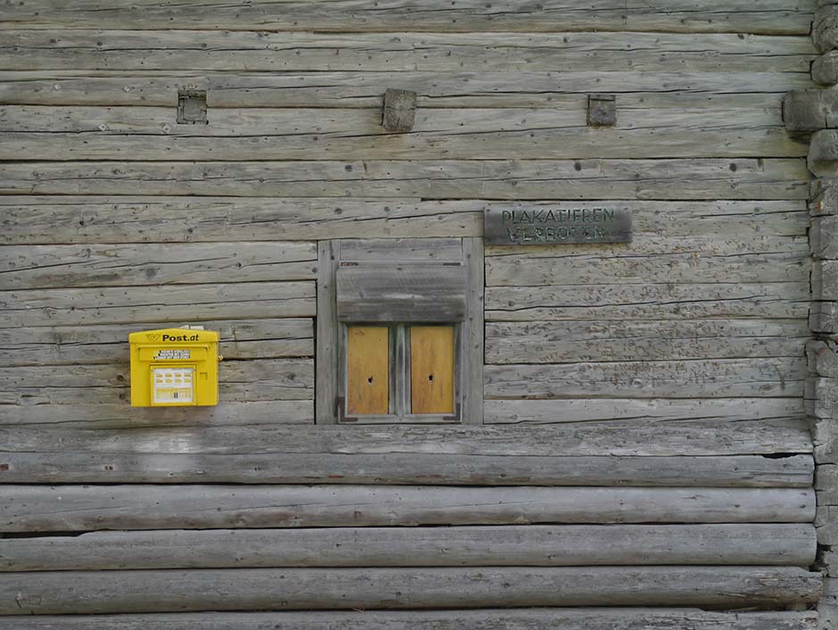 An der Wand eines alten Hauses im Lesachtal hängt ein gelber Briefkasten und ein Schild "Plakatieren verboten". Unter dem Schild sin die Abdrücke von Befestigungsnägeln.